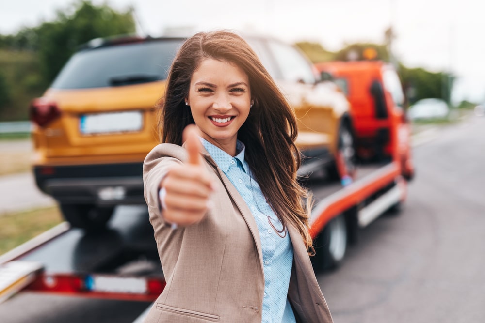 Woman Smiling And Showing Thumbs Up In Front Of A Car Being Towed